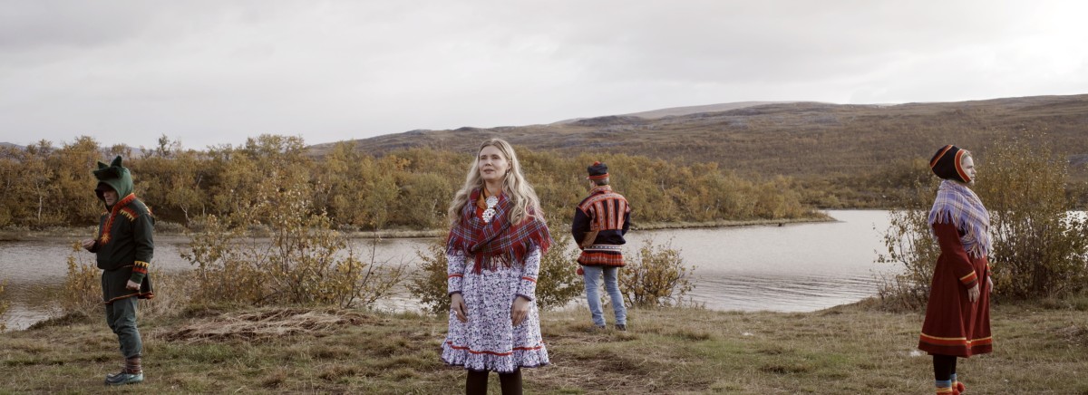 Bildet viser 4 personer med samiske folkedrakter som står med ryggen til hverandre ved en elv